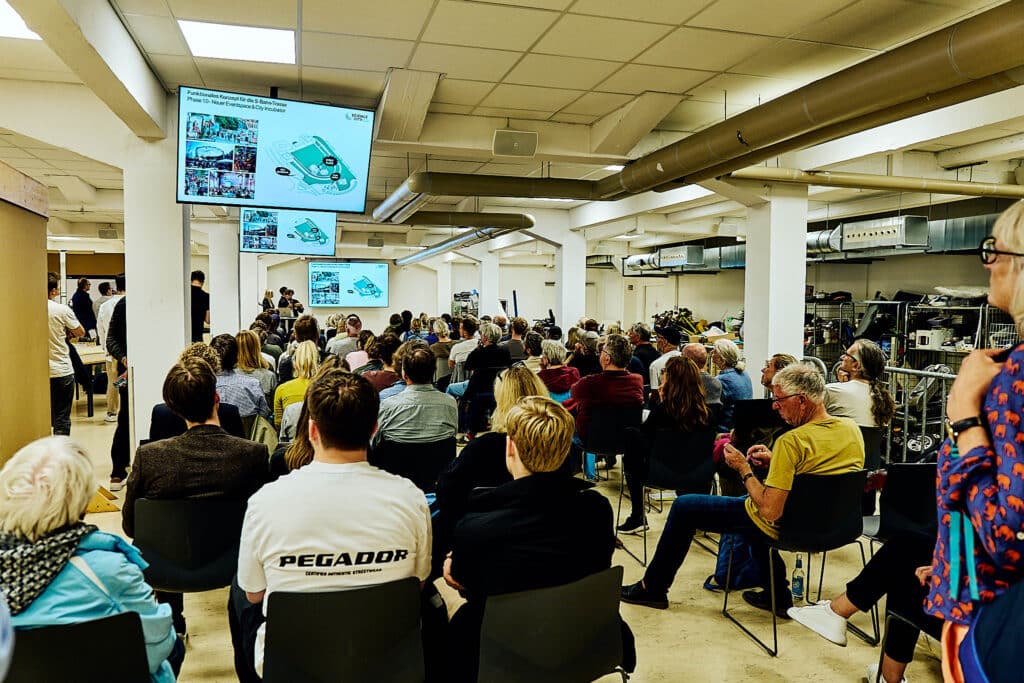 Mehrere Menschen sitzen in einem Raum auf Stühlen und interagieren miteinander. Dialog Quartiere am Volkspark 2024