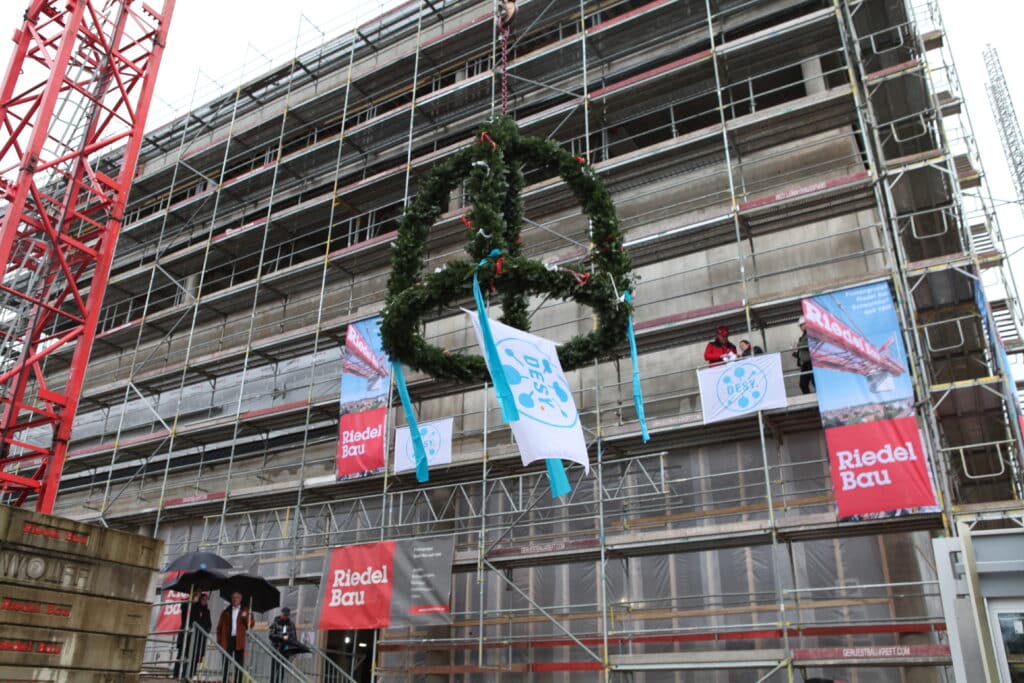 Ein im Bau befindliches Gebäude ist mit Baugerüsten umgeben, an denen mehrere "Riedel Bau"-Banner angebracht sind. Im Vordergrund wird ein Richtkranz mit grünen Zweigen und blauen Bändern von einem Kran hochgezogen. An dem Kranz ist eine Fahne mit dem Logo von "DESY" befestigt.