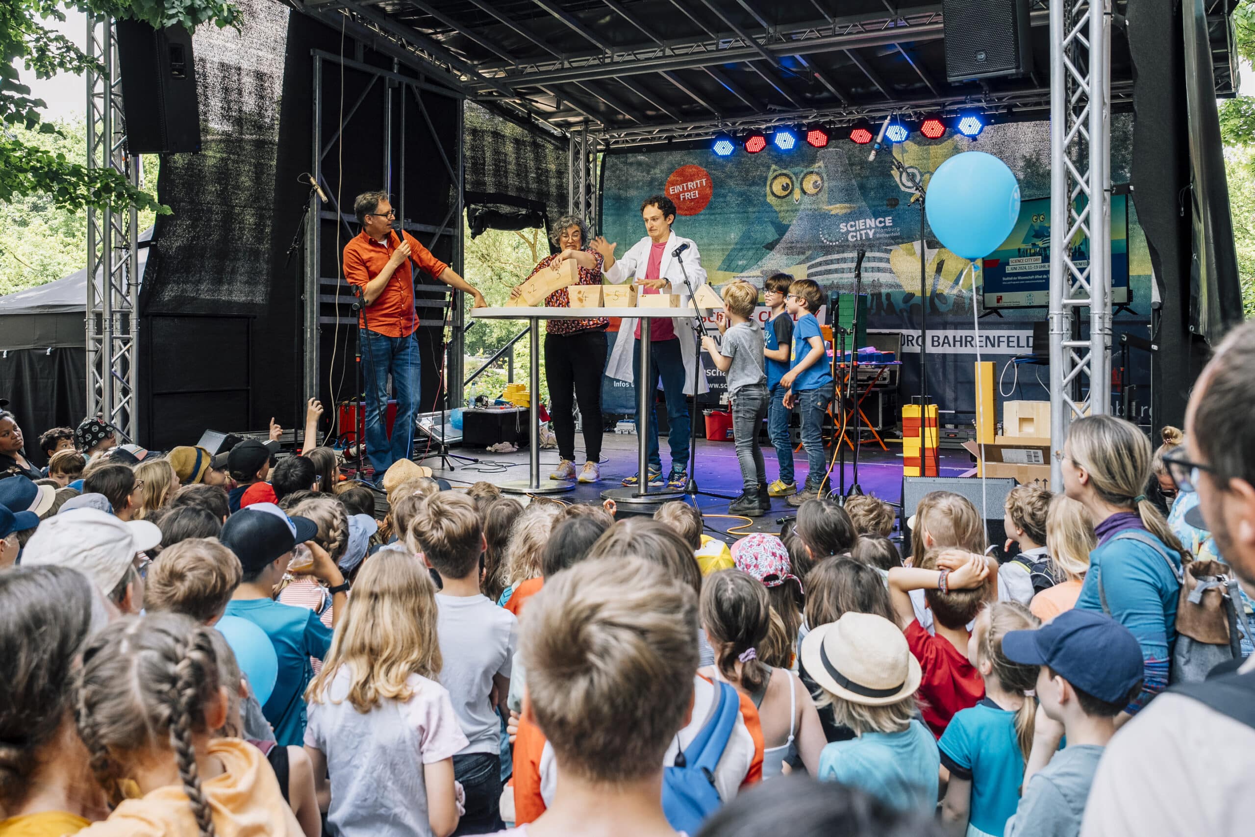 Eine Gruppe von Menschen, hauptsächlich Kinder, versammelt sich vor einer Bühne, auf der Erwachsene spielerisch mit anderen Kindern interagieren.