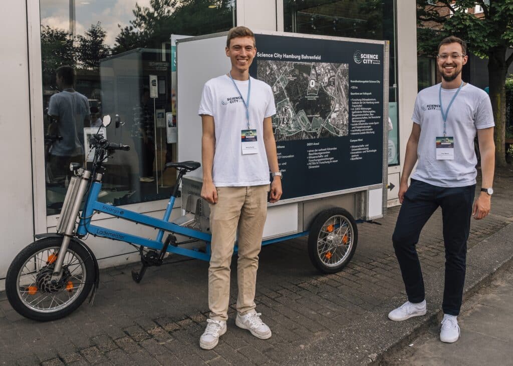 Zwei Männer mit T-Shirts der "Science City Hamburg Bahrenfeld" posieren vor einem Fahrrad im Rahmen der Science City Tour